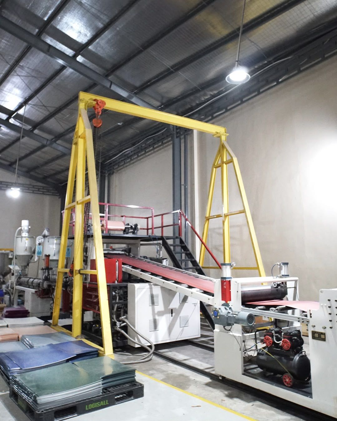 Lini mesin pembuat lembaran bahan di dalam pabrik, dengan katrol gantry kuning dan tumpukan panel berwarna di sampingnya.