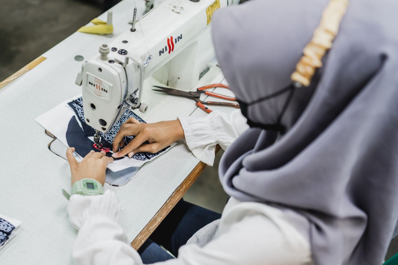 Detail dekat tangan penjahit menuntun bahan bermotif melewati jarum mesin jahit.