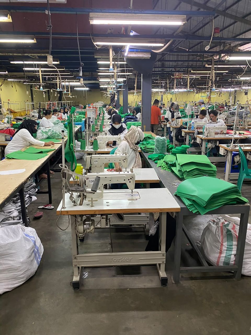 Lini jahit yang sedang berjalan, puluhan penjahit mengerjakan panel kain hijau di meja masing-masing.